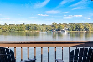 A lakeside view from a deck with chairs, a calm lake, and a distant house across the water. - Renovated River Home Hot Tub Sauna Game Room (Jeffersonville)