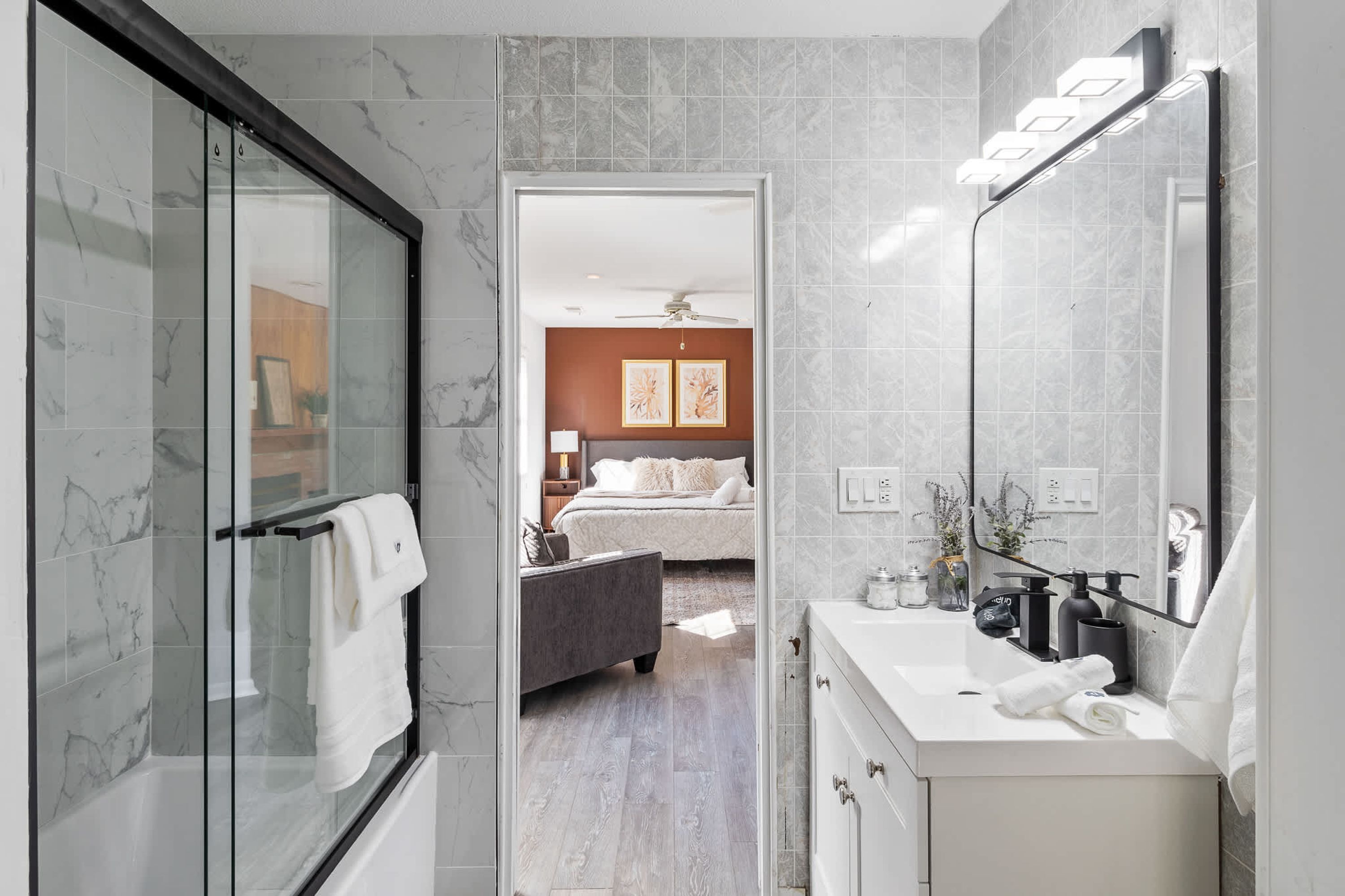 A modern bathroom with a glass shower enclosure, a white vanity with a mirror, and a visible bedroom with a bed and artwork.