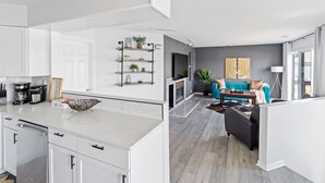 A modern kitchen with white cabinets, a marble countertop, and a coffee station. There is a living area with a blue sofa, a fireplace, and a coffee table. - Renovated River Home Hot Tub Sauna Game Room (Jeffersonville)