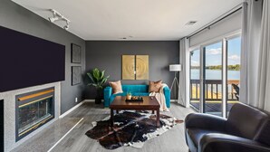 A modern living room with a fireplace, a blue sofa, a coffee table, and a view of a lake through large windows. - Renovated River Home Hot Tub Sauna Game Room (Jeffersonville)