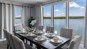 A dining area with a view of a lake, a table set with white dinnerware, and grey upholstered chairs. - Renovated River Home Hot Tub Sauna Game Room (Jeffersonville)