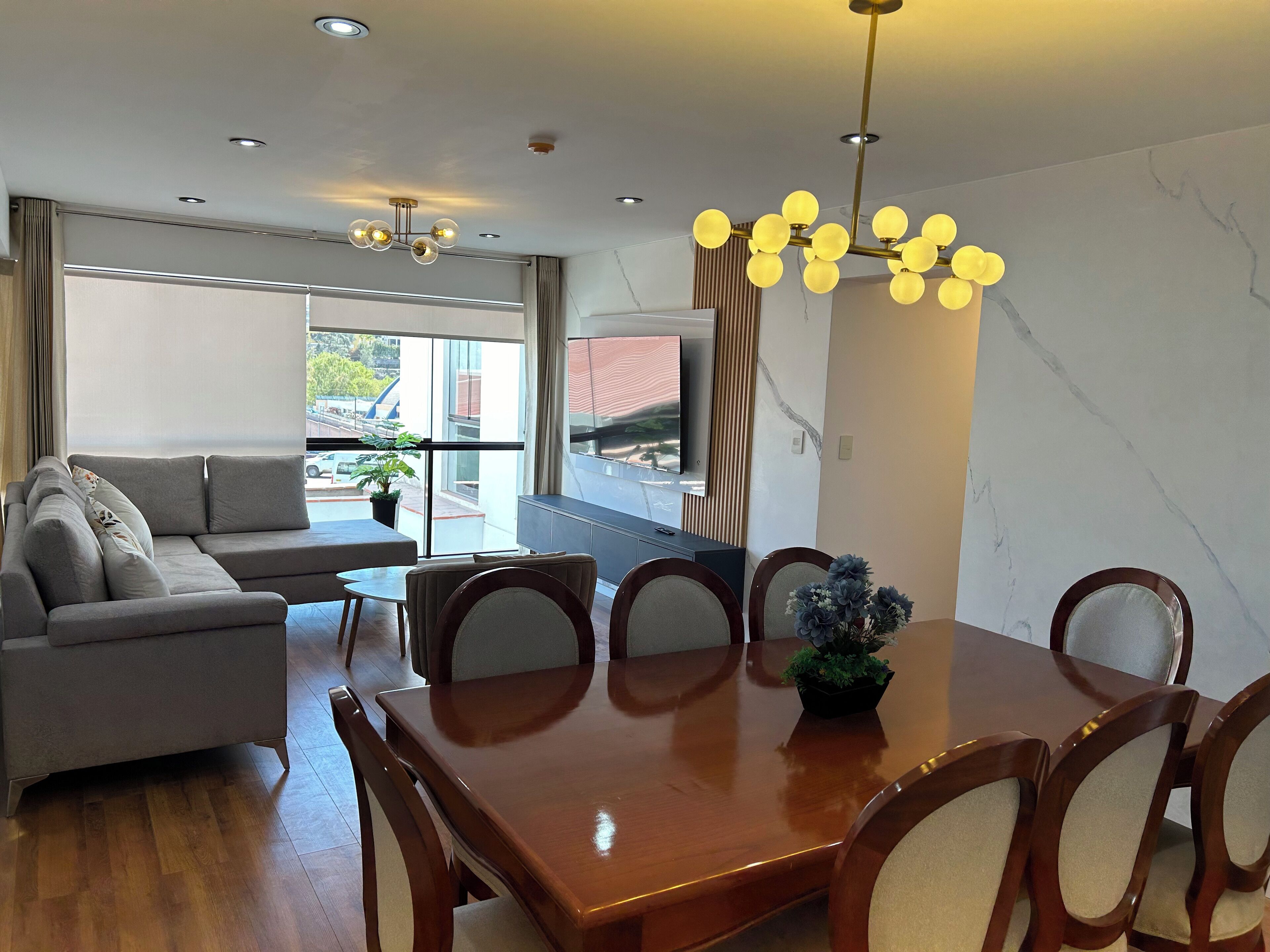 Un salon moderne avec un canapé, une table à manger et des chaises. Un téléviseur est fixé au mur, et une fenêtre offre une vue sur l’extérieur.