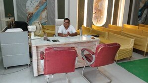 A hotel reception area with a marble counter, red chairs, a fan, and yellow cushioned seating.