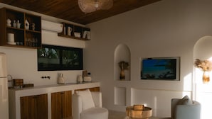 A modern kitchen with white cabinetry, a built-in oven, and a large island with a sink. There are two white armchairs and a round wooden coffee table.