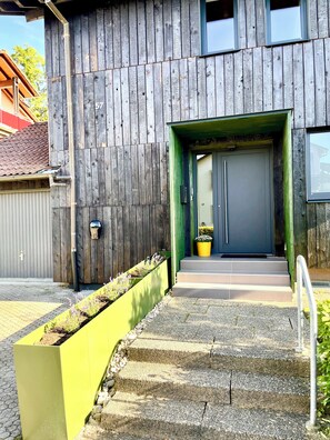 Um edifício moderno com fachada de madeira, entrada verde e um conjunto de degraus de concreto que levam a uma porta.
