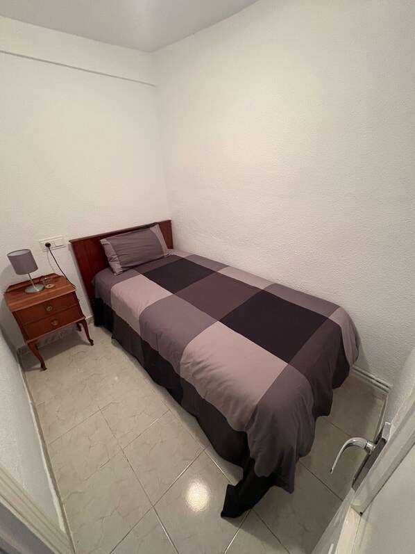 A small, single-bed room with a wooden headboard, a bedside table with a lamp, and a tiled floor.