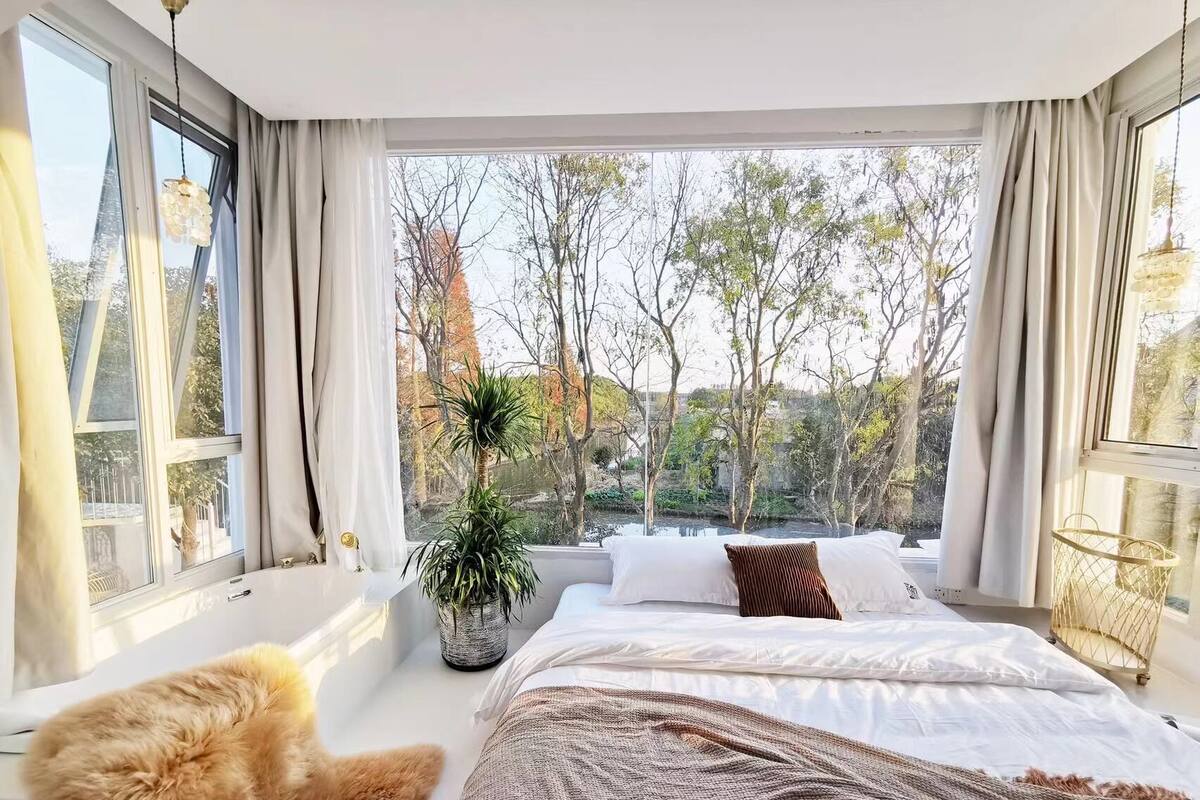 Un dormitorio con un ventanal grande, una cama con ropa de cama blanca, una manta marrón y una planta en maceta.