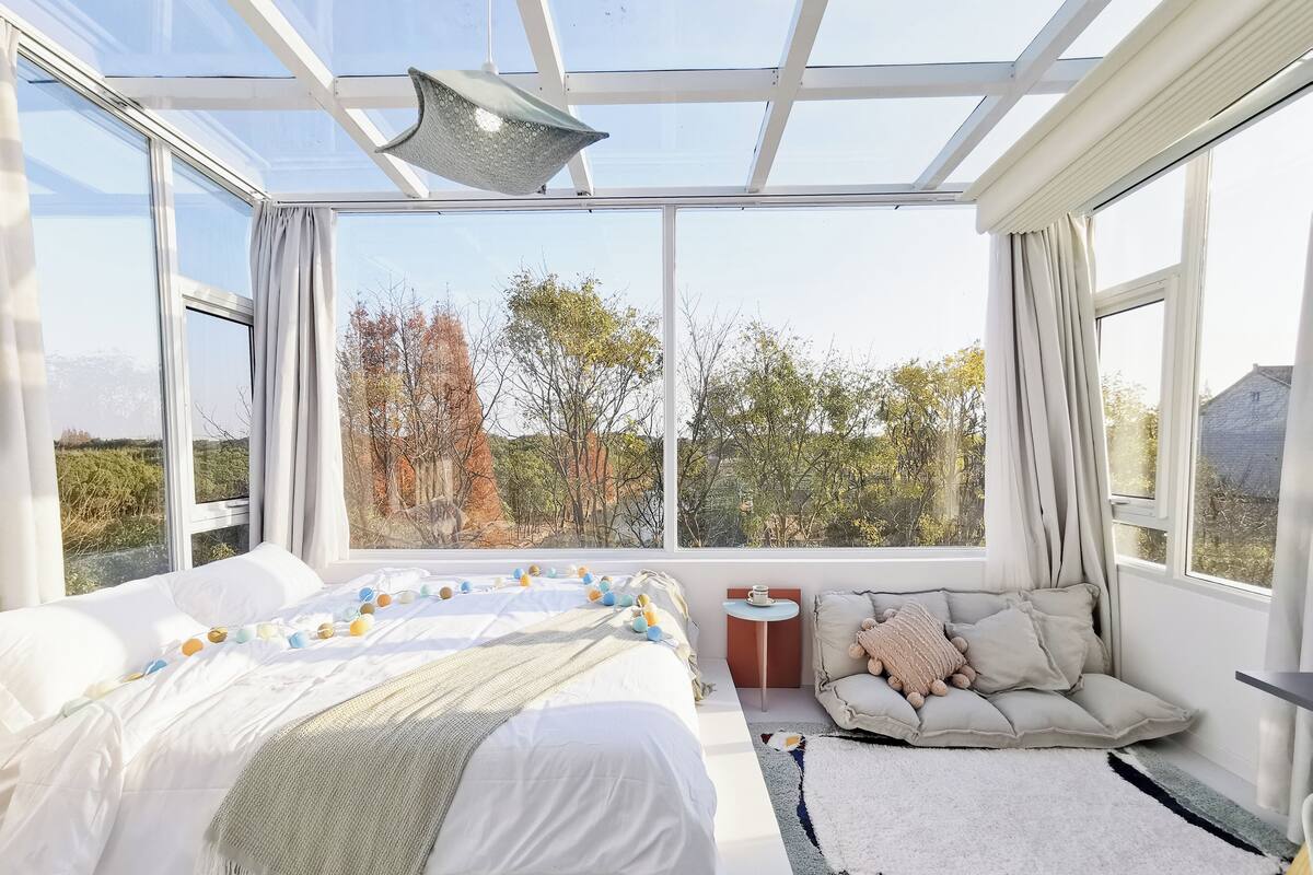 Un dormitorio con una cama grande, un sofá, una mesita y vistas a árboles a través de amplios ventanales.
