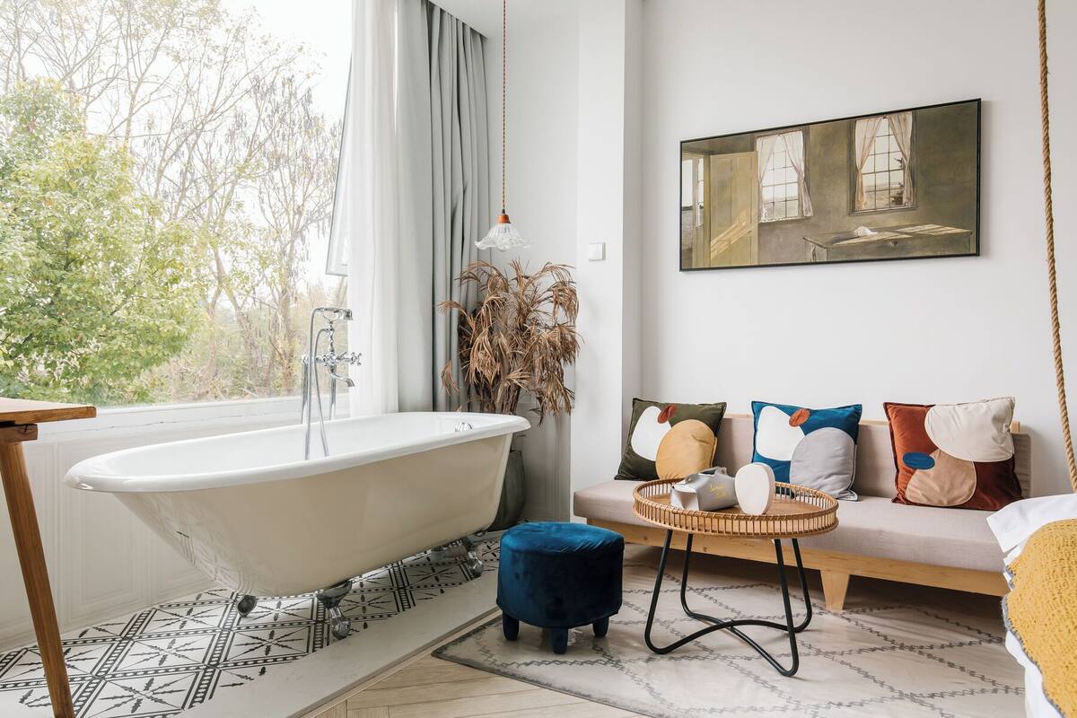 Un baño con bañera independiente, una ventana con cortinas y vistas a árboles.