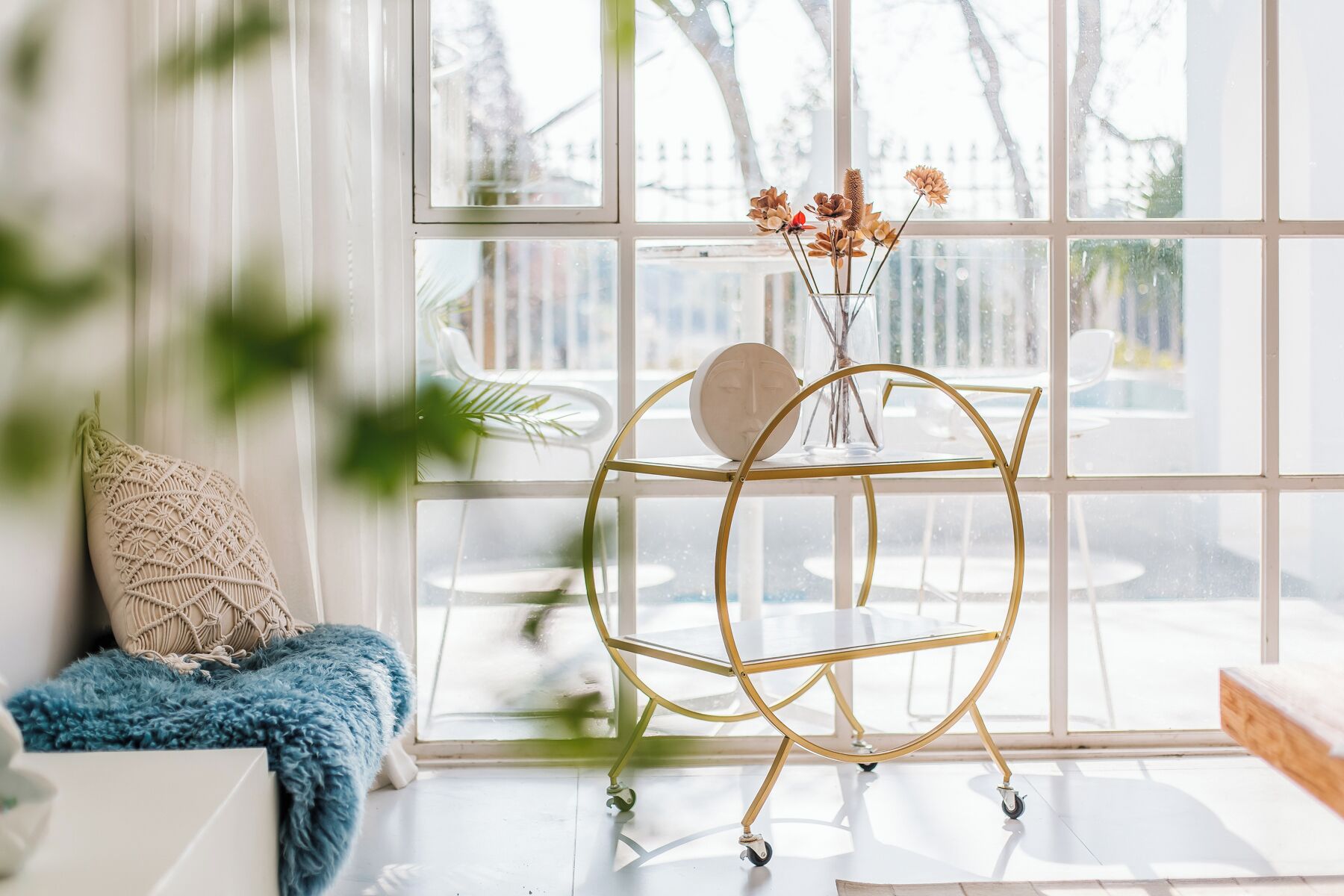 A room with a large window, a gold-framed cart, a macrame cushion, and a blue throw blanket.