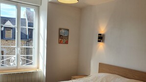 A bedroom with a bed, a window, a wall-mounted light, and a radiator. - Fully furnished and equipped apartment in the courtyard of the Haut Manoir (Villedieu-les-Poêles)