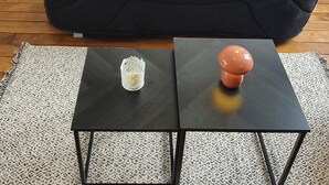 A black sofa with fluffy white and red pillows, a small table with a decorative item, and a patterned rug. - Fully furnished and equipped apartment in the courtyard of the Haut Manoir (Villedieu-les-Poêles)