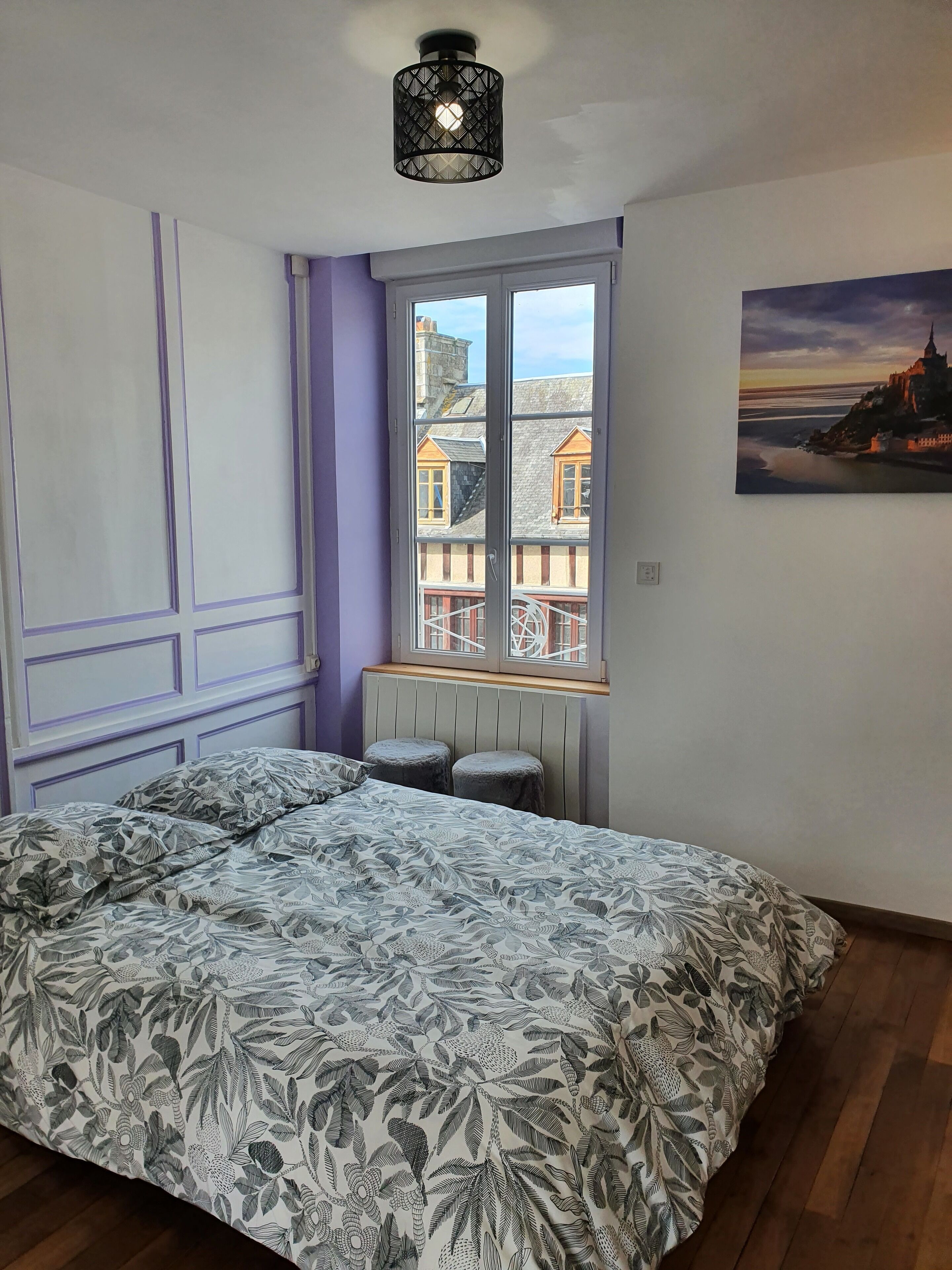 Un dormitorio con cama, ventana con vista a edificios y un cuadro en la pared.