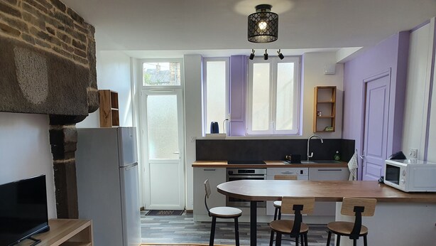 A compact kitchen with a stone wall, a wooden dining table, and a TV on a shelf. - Fully furnished and equipped apartment in the courtyard of the Haut Manoir (Villedieu-les-Poêles)