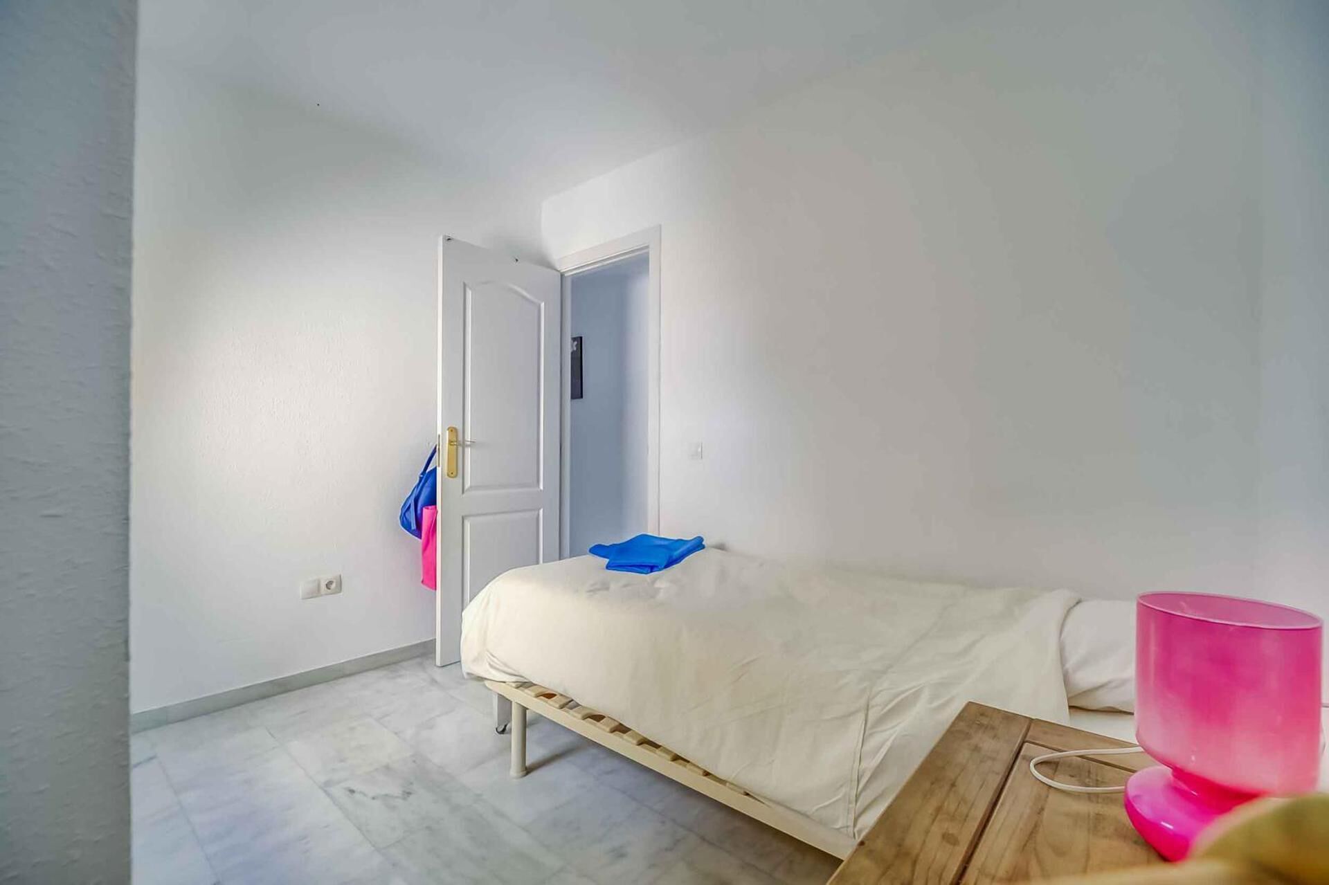A room with a bed, a wooden bench, a pink lamp, and a door leading to another room.