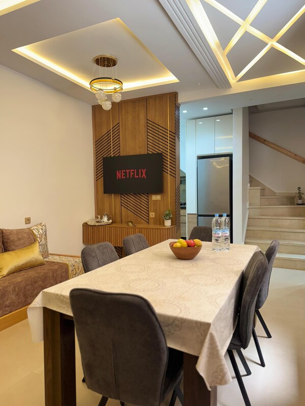 A modern living room with a dining area, a flat-screen TV displaying 'NETFLIX', and a staircase leading to another level.