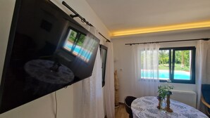 Una habitación con un televisor de pantalla grande montado en la pared, una mesa redonda con mantel floreado y una ventana con vistas a una piscina.