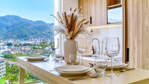 Ein Esstisch ist gedeckt mit Tellern, Gläsern und Besteck, in der Mitte steht eine Vase mit getrocknetem Pampasgras. Man blickt auf eine Stadtlandschaft und Berge.