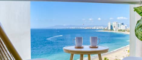 A balcony with a view of the ocean, two white chairs, and a small round table with two white cups.