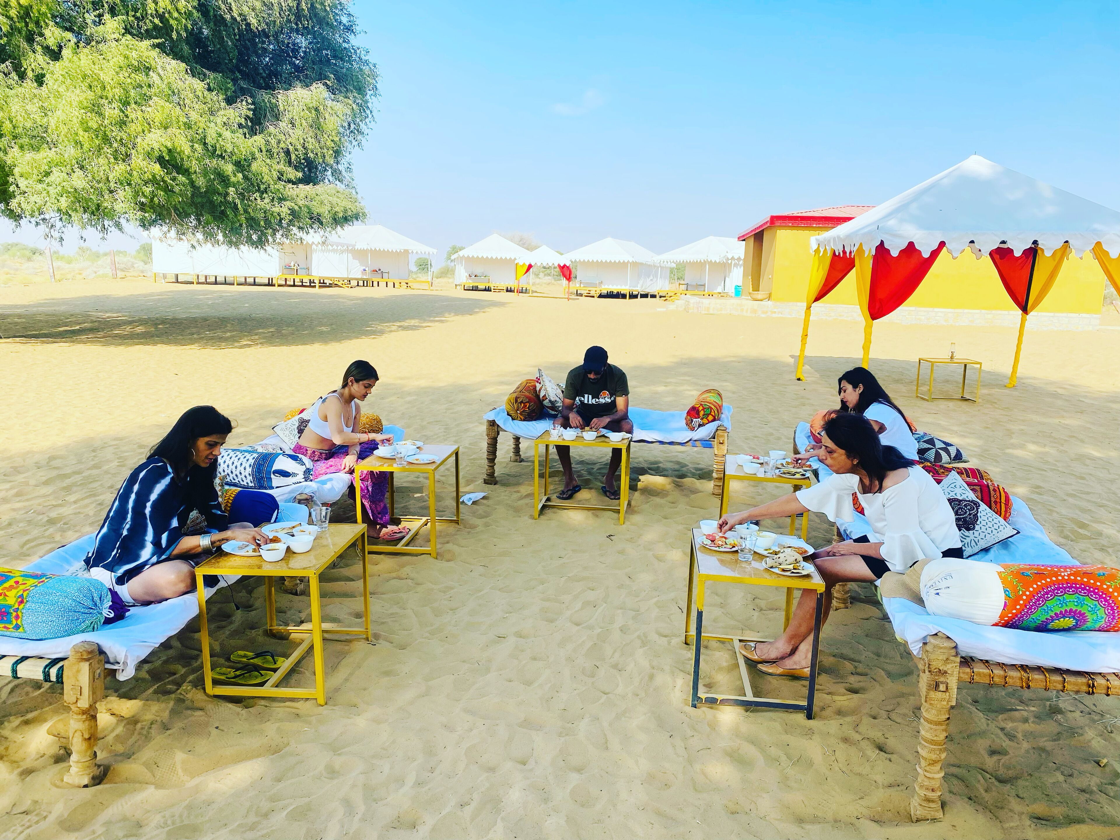 A group of people dining at tables on a sandy beach with tents in the background.