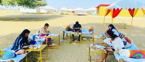 A group of people dining at tables on a sandy beach with tents in the background.