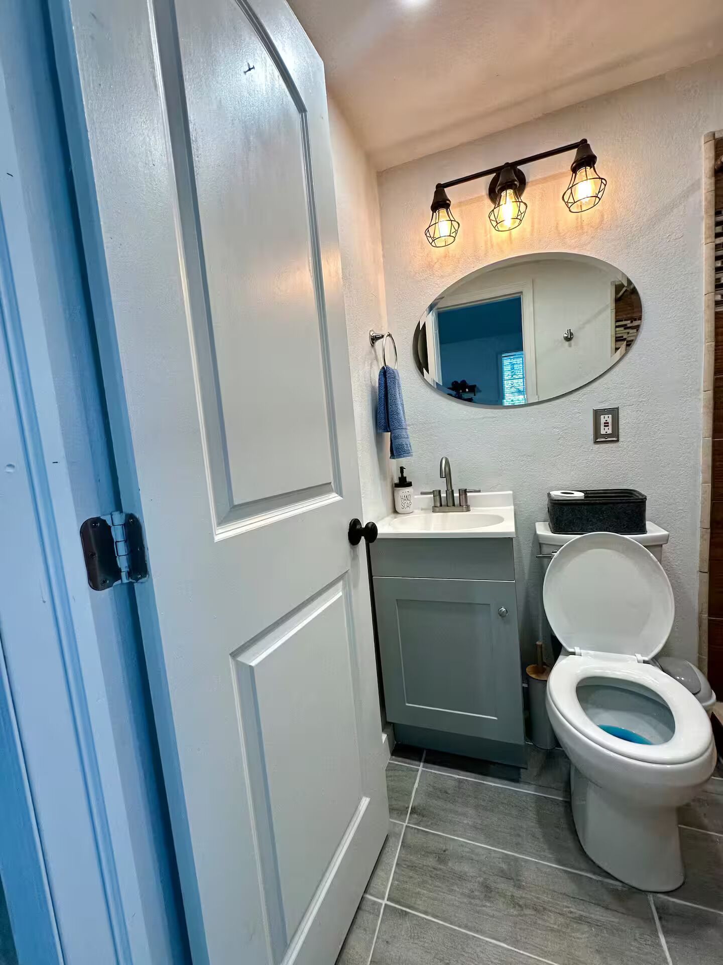 Un baño con puerta blanca, un lavamanos con espejo, un inodoro y una toalla colgada en un perchero.