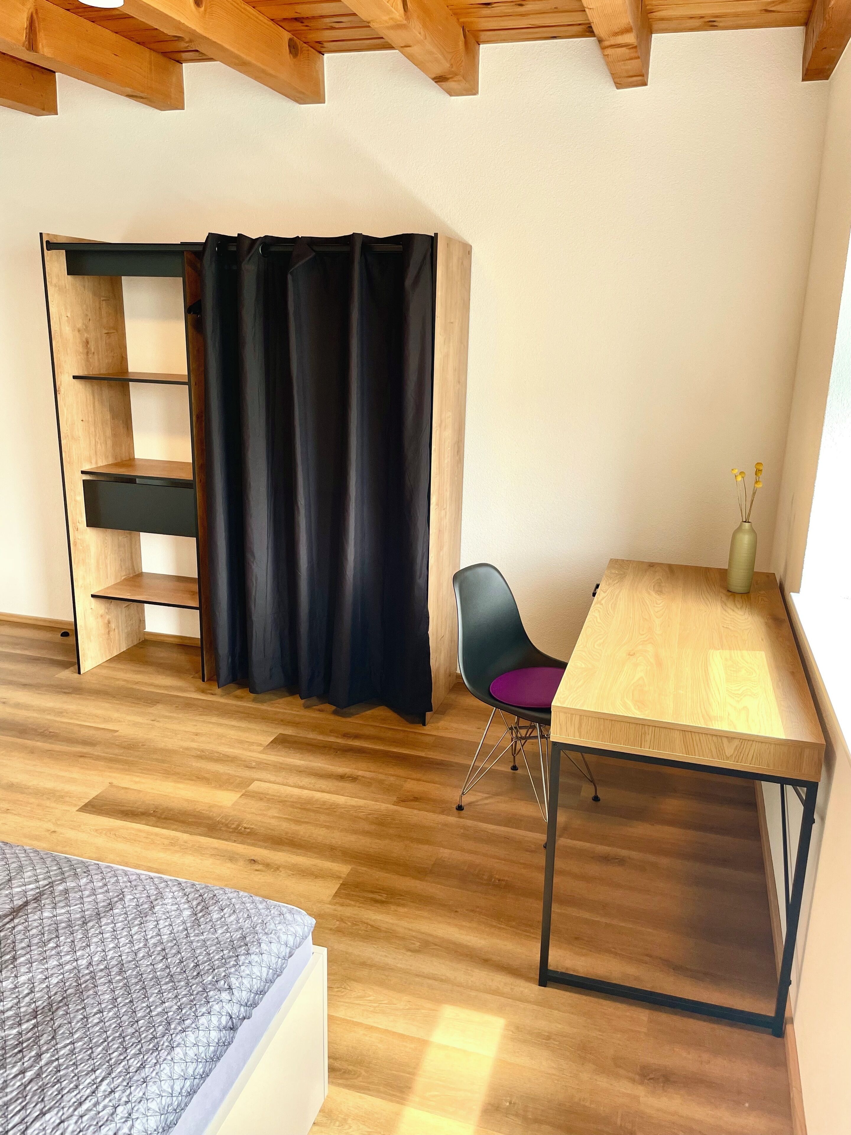 Ein Zimmer mit Bett, Schreibtisch, Stuhl, einer Holzregaleinheit und einem schwarzen Vorhang.
