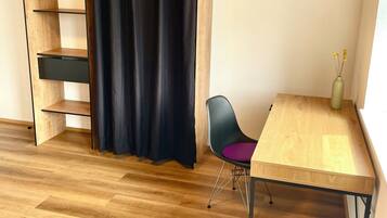 Ein Zimmer mit Bett, Schreibtisch, Stuhl, einer Holzregaleinheit und einem schwarzen Vorhang.