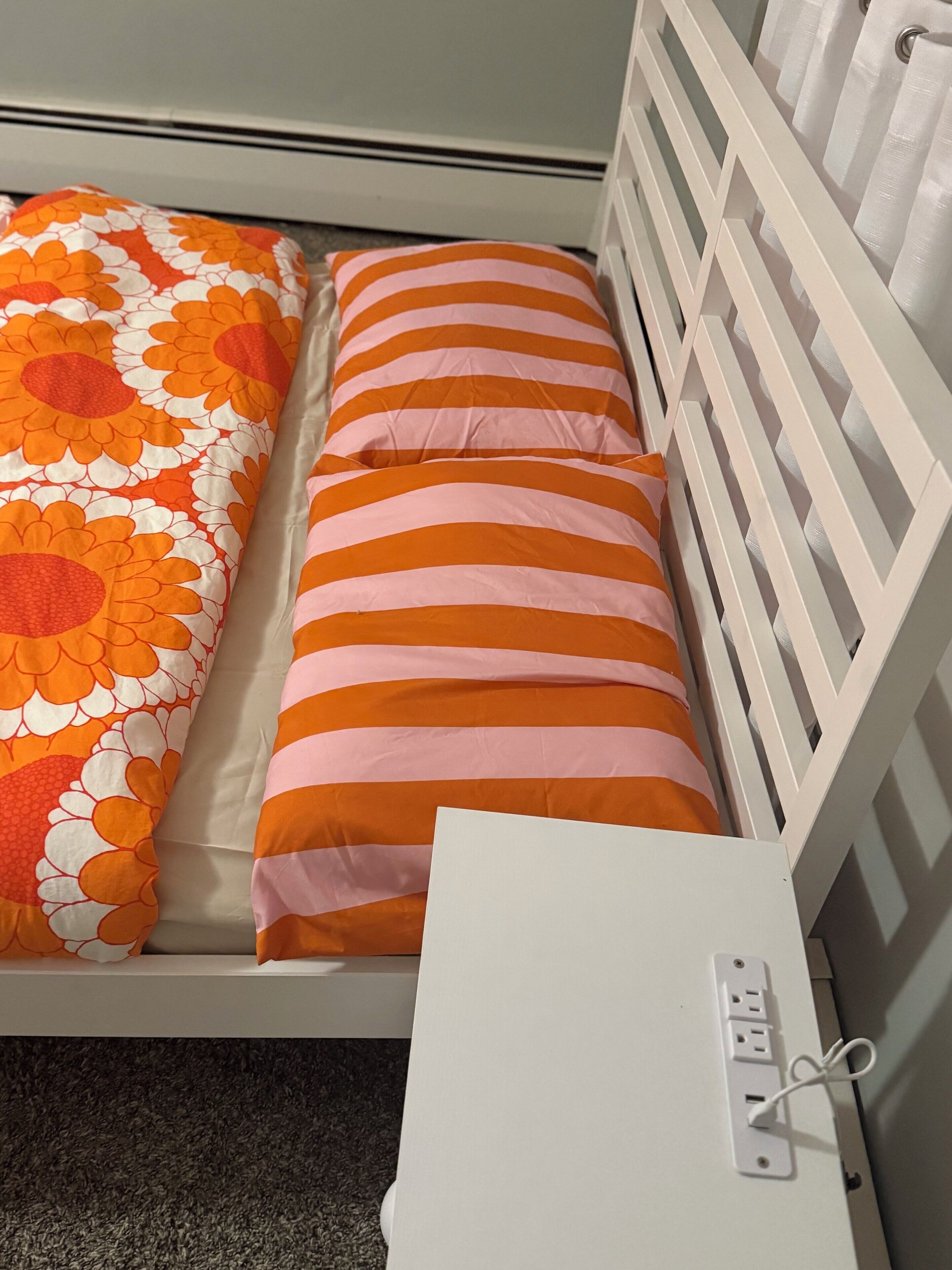 Ein Bett mit einer floralen Decke und gestreiften Kissen, ein weißer Kopfteil und eine Steckdose an der Seite.