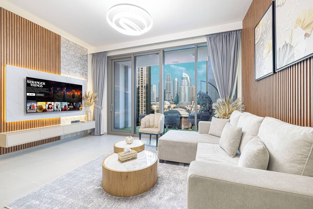 A modern living room with a flat-screen TV, a round coffee table, a beige sofa, and a city view through large windows.