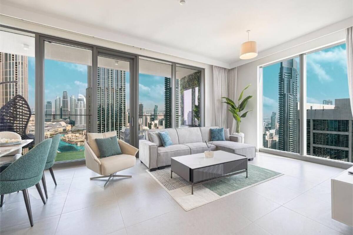 A modern living room with a sofa, coffee table, and chairs, large windows with a city view, and a dining area with a table and chairs.