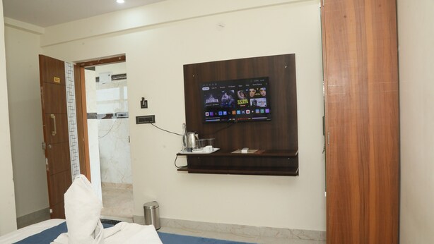 A hotel room with a bed, a TV mounted on the wall, a small shelf with toiletries, and a wooden door. - Hotel Rest O ITPL (Bangalore)