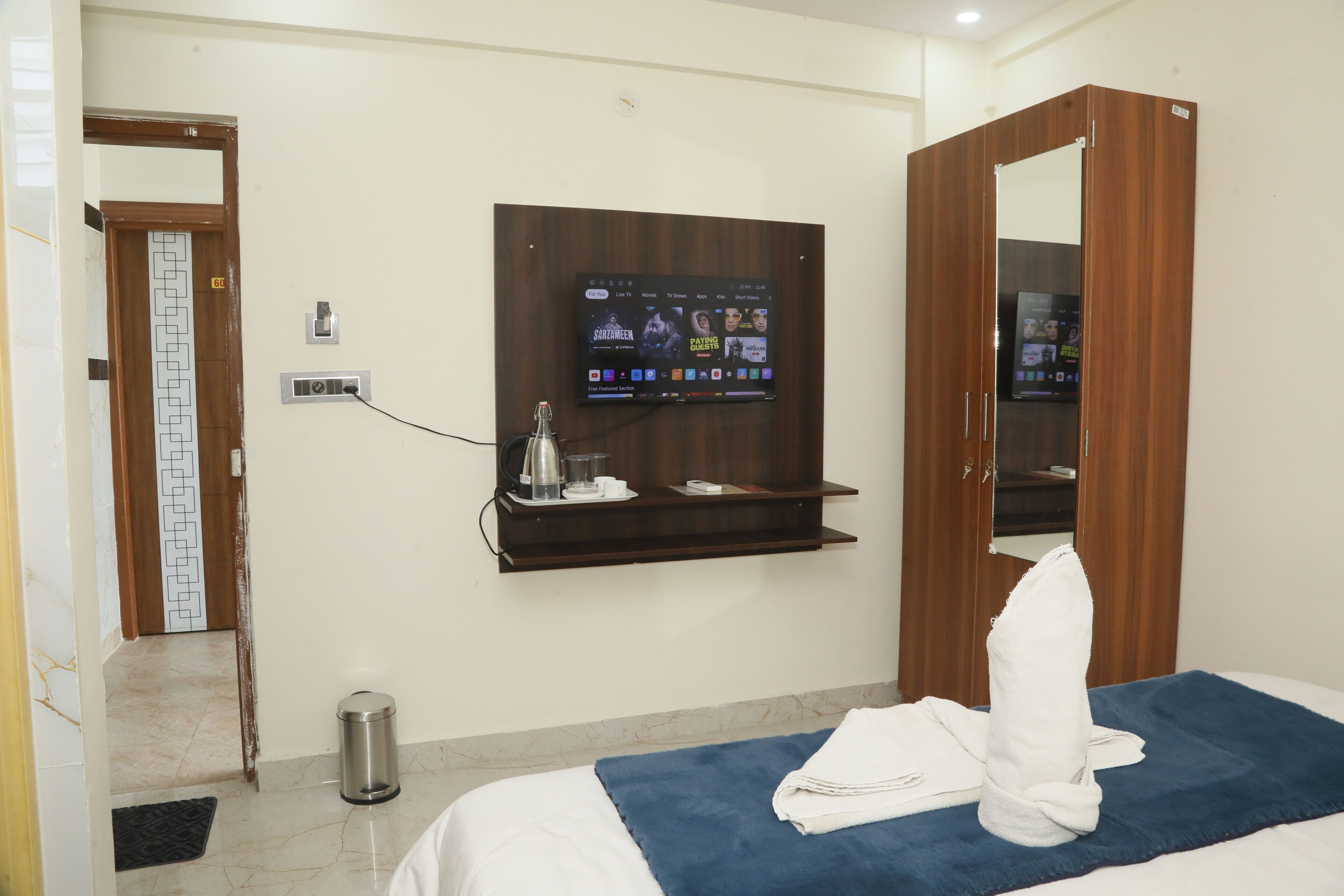 A hotel room with a bed, a TV mounted on the wall, a wooden wardrobe, and a small shelf with a kettle and cups.