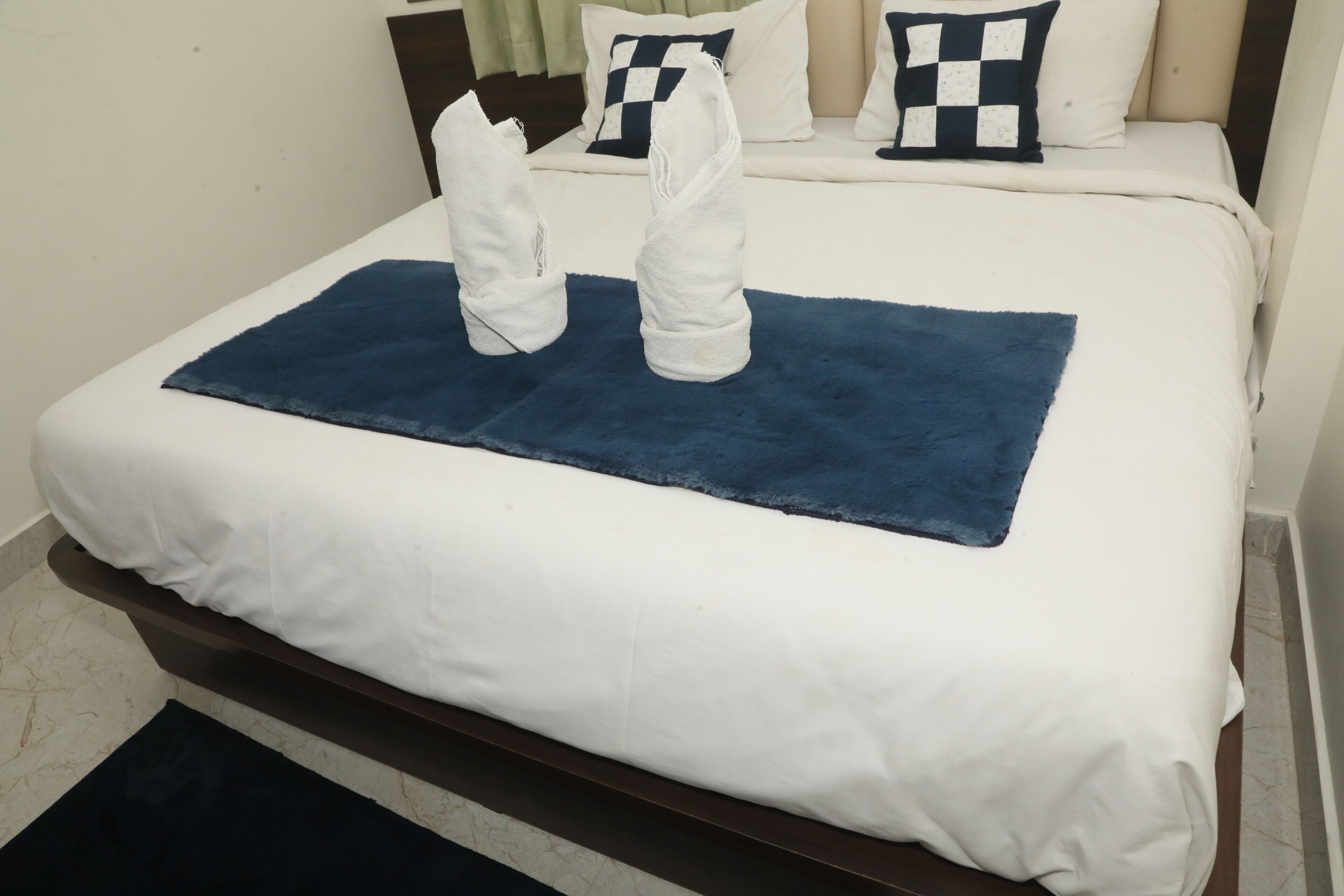 Un lit bien fait, avec une literie blanche et un tapis bleu en guise d’élément décoratif, deux serviettes pliées posées sur le lit, et des coussins à carreaux sur la tête de lit.