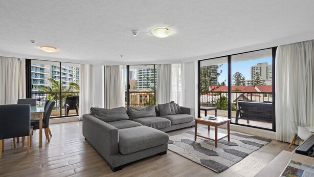 A spacious living room with a grey sectional sofa, a dining area with a table and chairs, large windows offering views of buildings and palm trees. - Stylish 3BRM Haven in Surfers Paradise (Surfers Paradise)