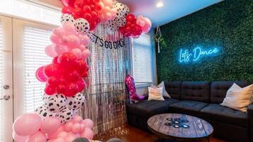 A room with a neon sign reading "Let's Dance," a sofa, a round wooden table, and a decorative balloon arch with the text "It's Go Girls."