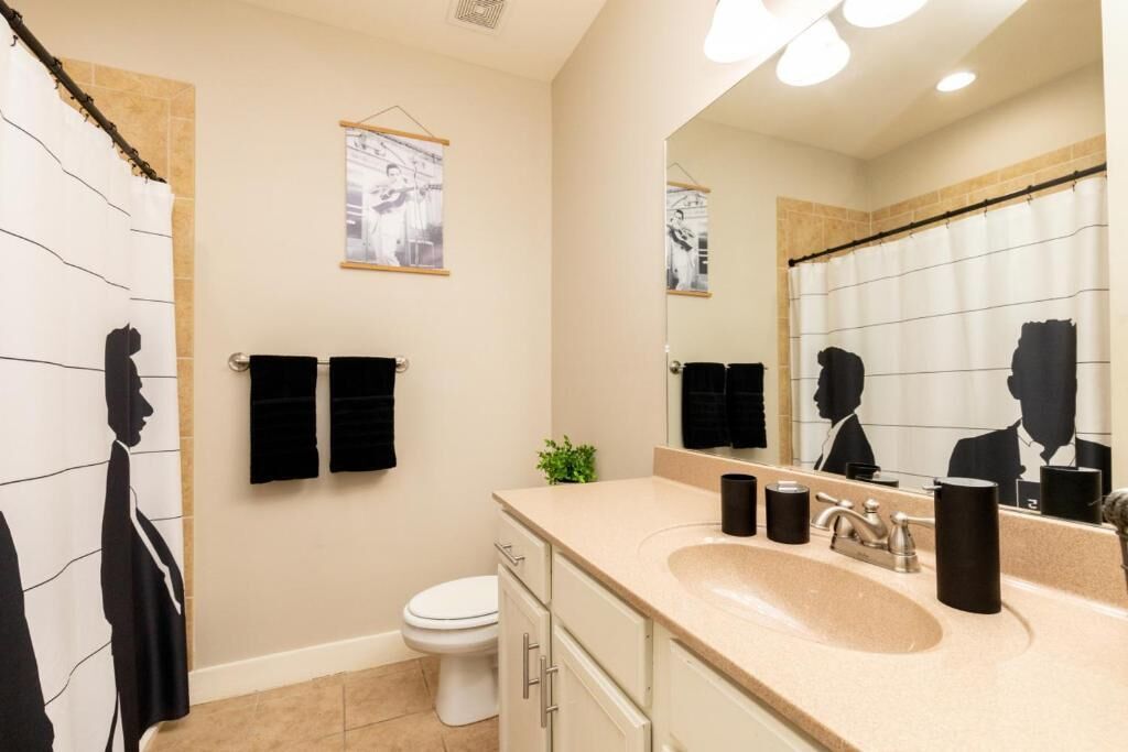 Une salle de bain moderne avec un mur recouvert de carreaux beiges, un grand miroir, un lavabo doté d’un plan de toilette beige et deux serviettes noires suspendues à des crochets.