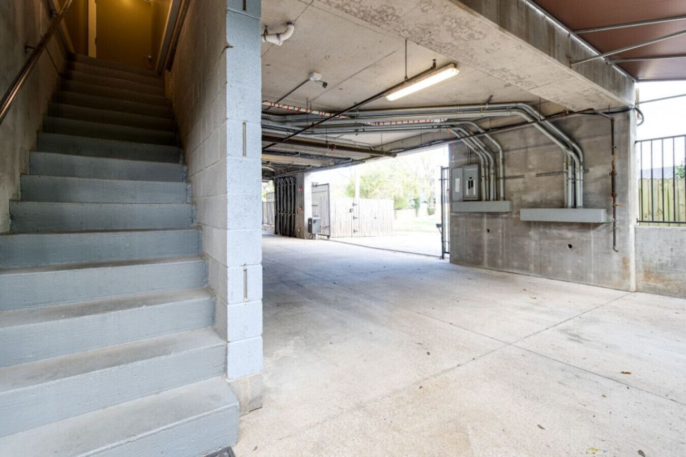 A concrete stairway leading to a covered parking garage with visible pipes and a metal railing.