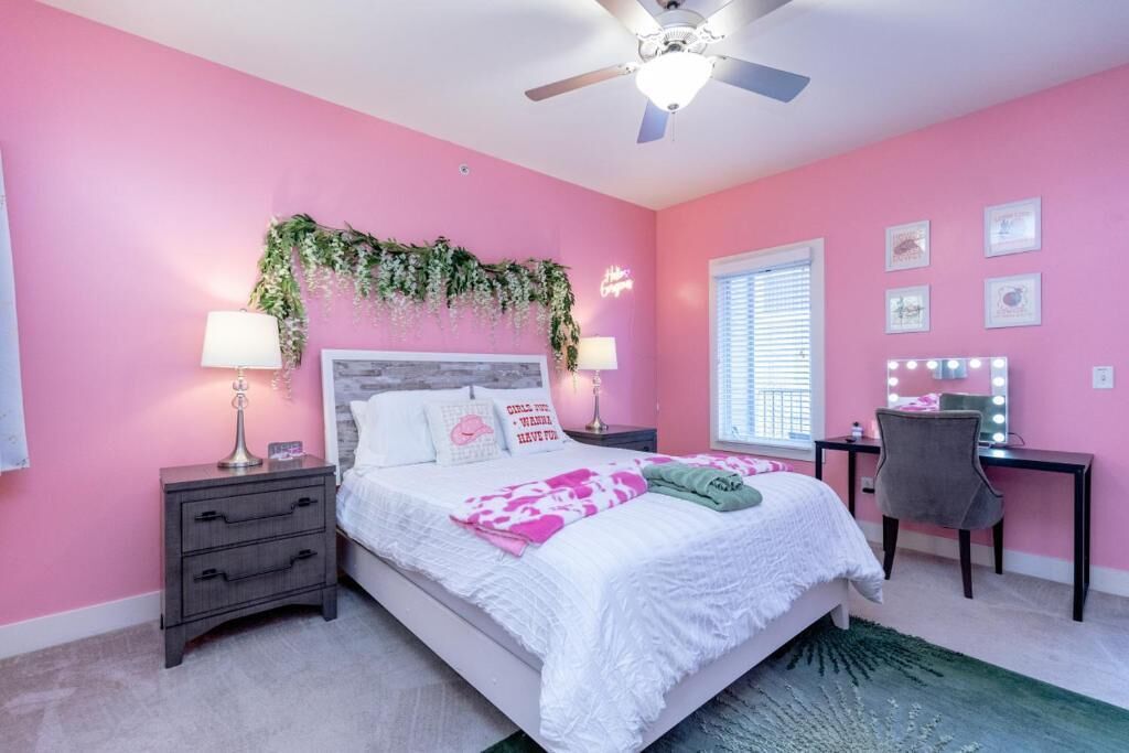 A pink-themed bedroom with a bed, bedside tables, a desk with a chair, a ceiling fan, and a window with blinds.
