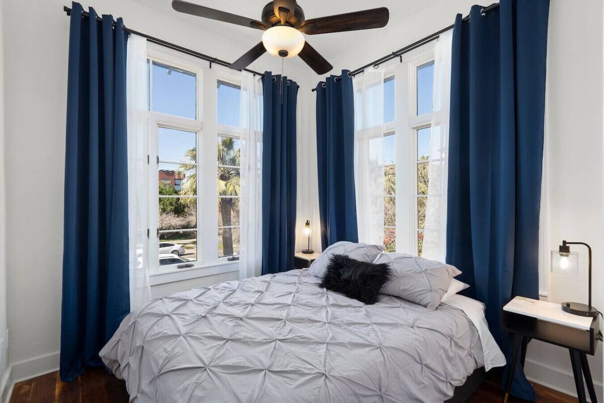 Quarto com cama, ventilador de teto e vista para o exterior através de grandes janelas.