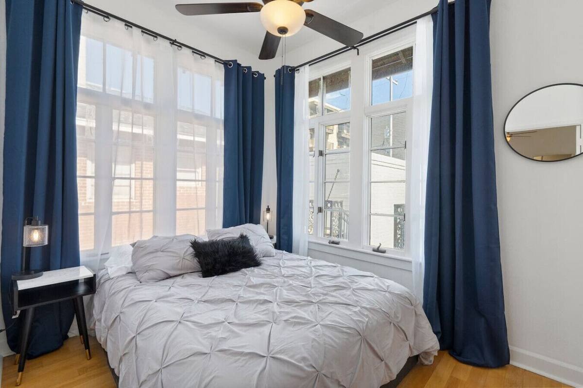 Quarto com cama, escrivaninha, cadeira, ventoinha no teto e janela com cortinas.