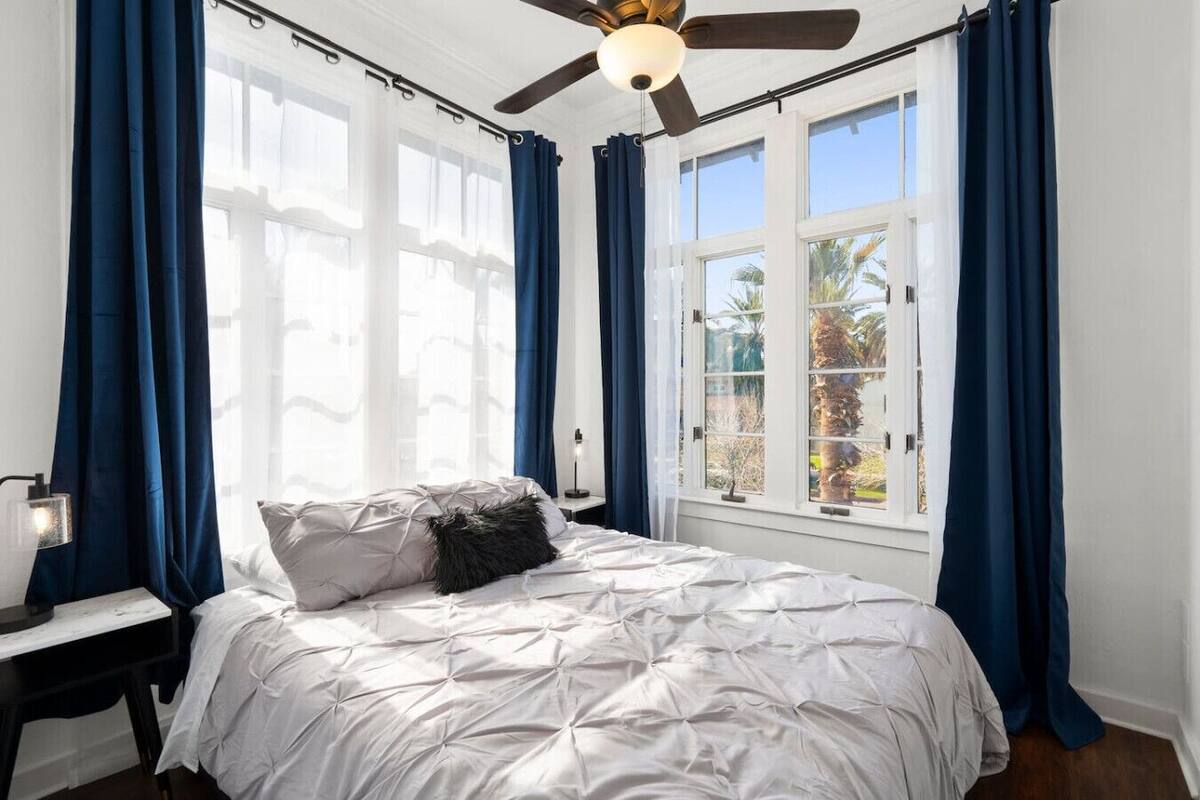 Quarto com cama, ventilador de teto e janelas amplas com cortinas.
