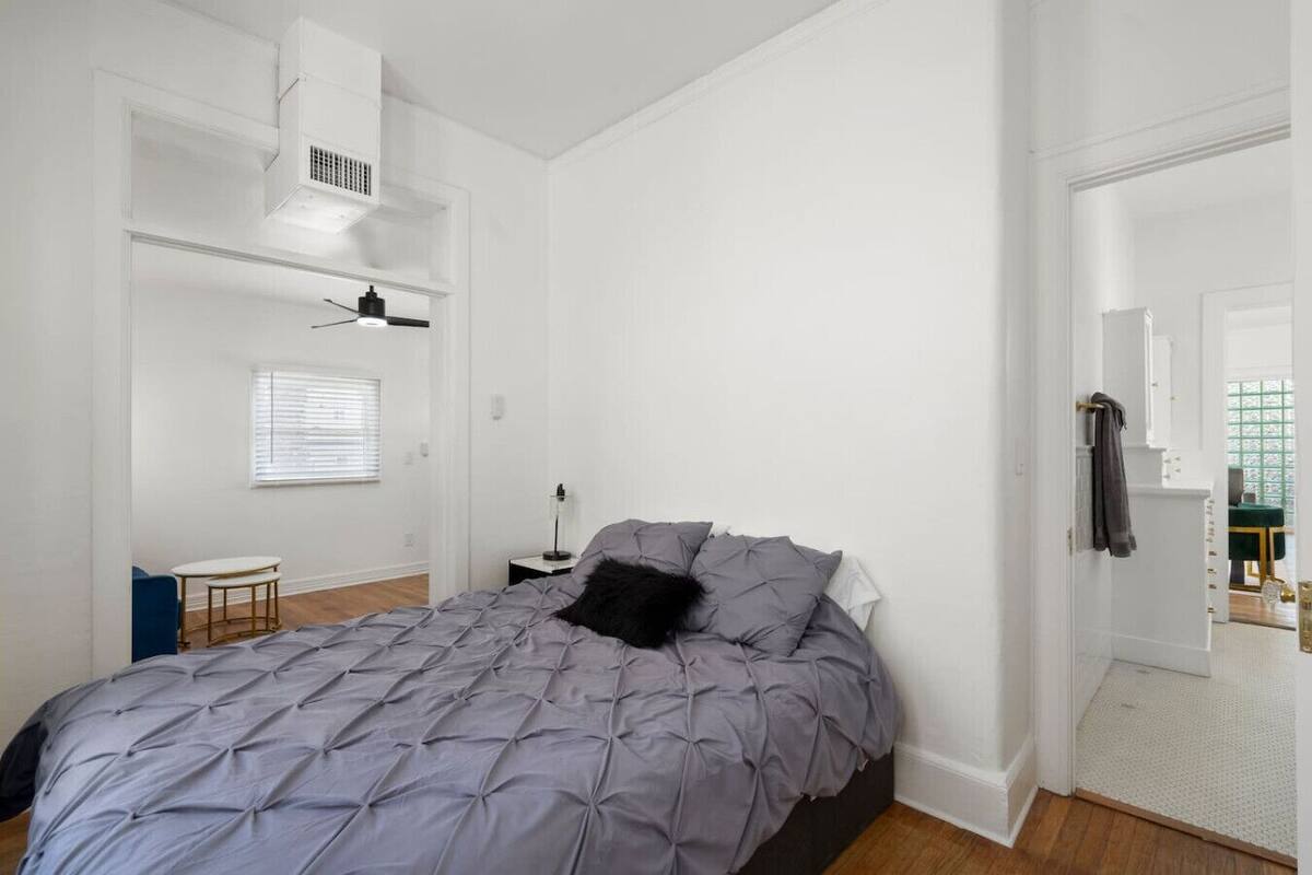 Quarto com cama, uma mesinha e uma cadeira. Há uma janela com persianas, um ventilador de teto e uma porta que dá para outro cômodo.