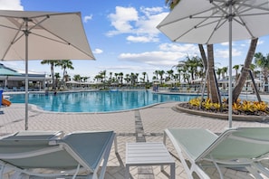A poolside area with lounge chairs and umbrellas, surrounded by palm trees and a clear sky.