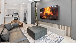 A modern living room with a grey sofa, a grey ottoman, and a large flat-screen TV mounted on the wall displaying a fireplace.