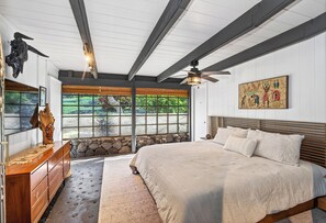 Un dormitorio con una cama grande, cabecera de madera, ventilador de techo, pared de piedra y una ventana con vistas a la vegetación.