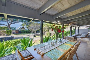 Un espacio de comedor al aire libre techado con una mesa para dos, rodeado de abundante vegetación y vistas a una calle residencial.
