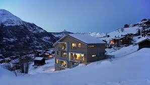 Um chalé de dois andares com varanda, rodeado por montanhas cobertas de neve e outros edifícios.