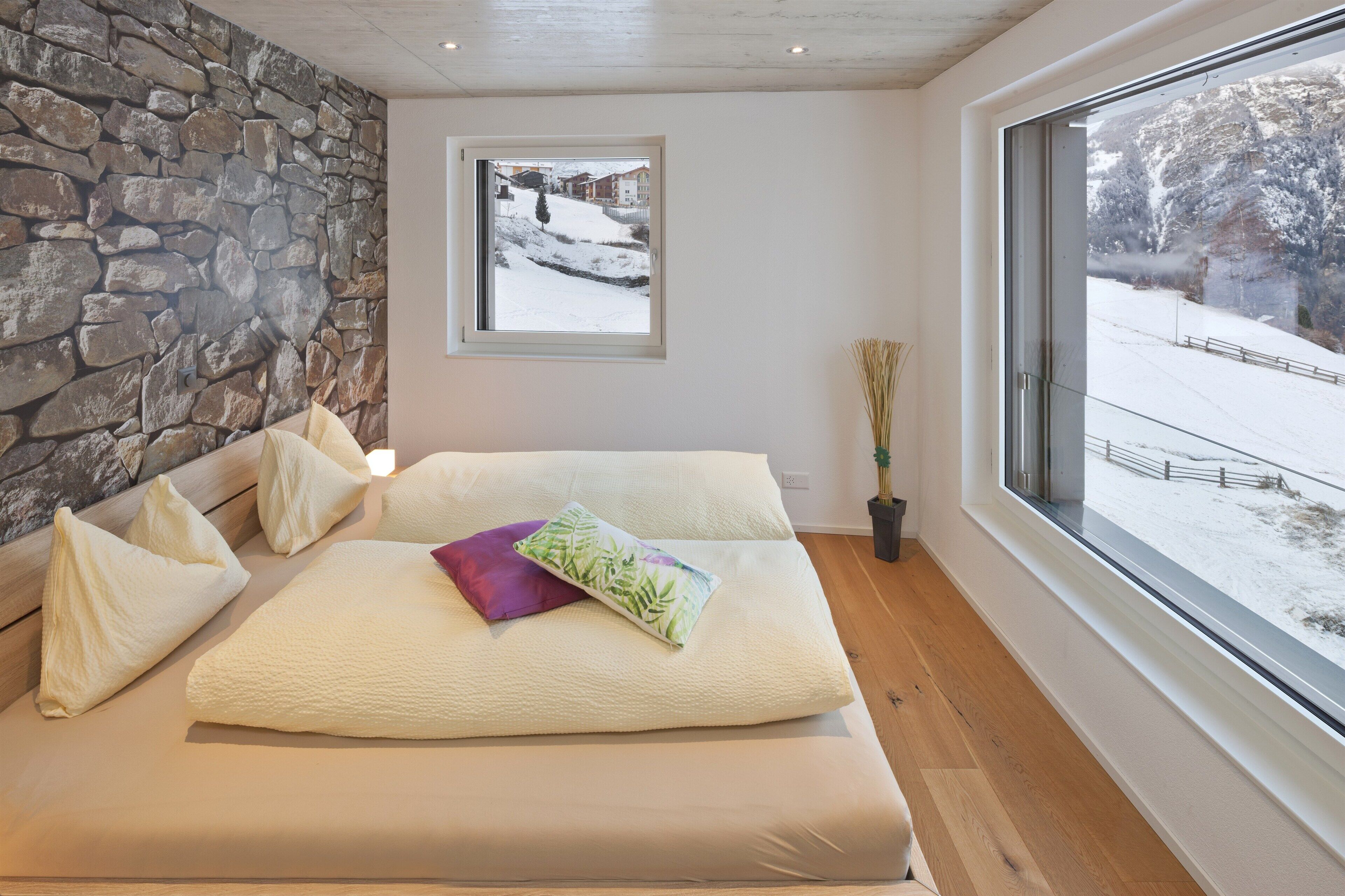 A bedroom with a stone wall, a large window with a snowy view, and a bed with pillows.