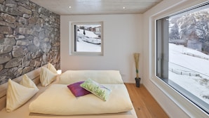 Quarto com parede de pedra, uma janela grande com vista nevada e uma cama com almofadas.